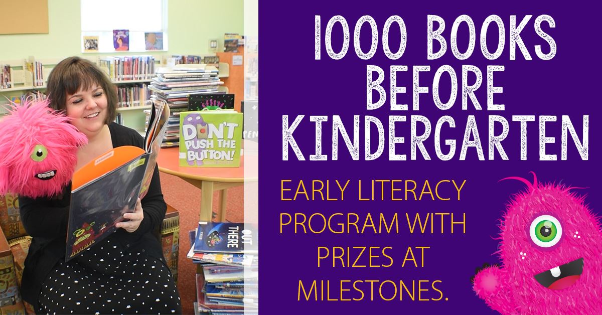 Library staff reading book to pink monster mascot next to stacks and stacks of books