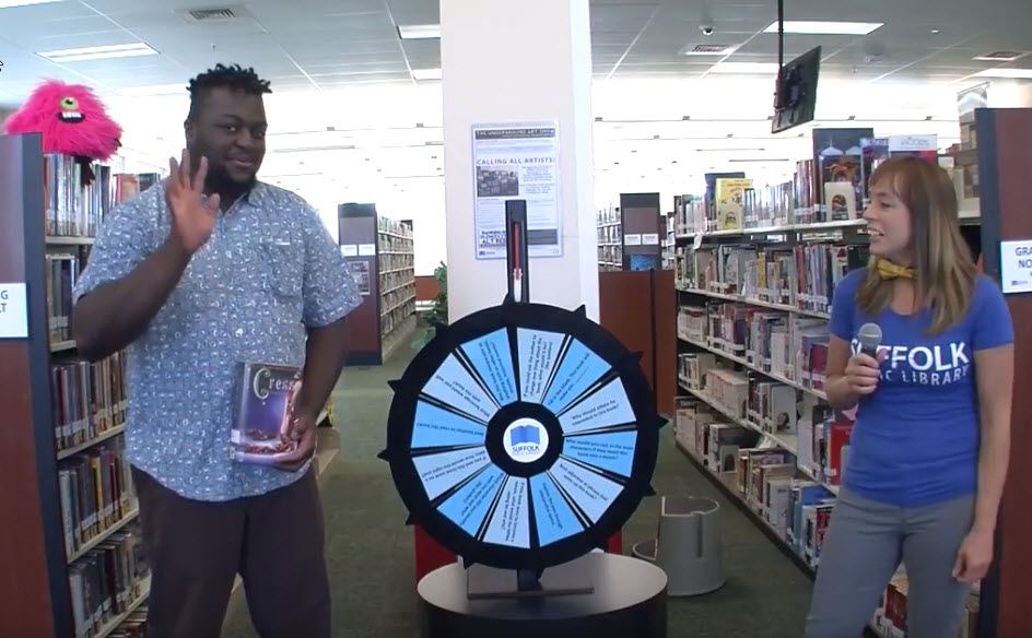 Still shot of Face the Wheel video of man holding book next to a wheel and female host